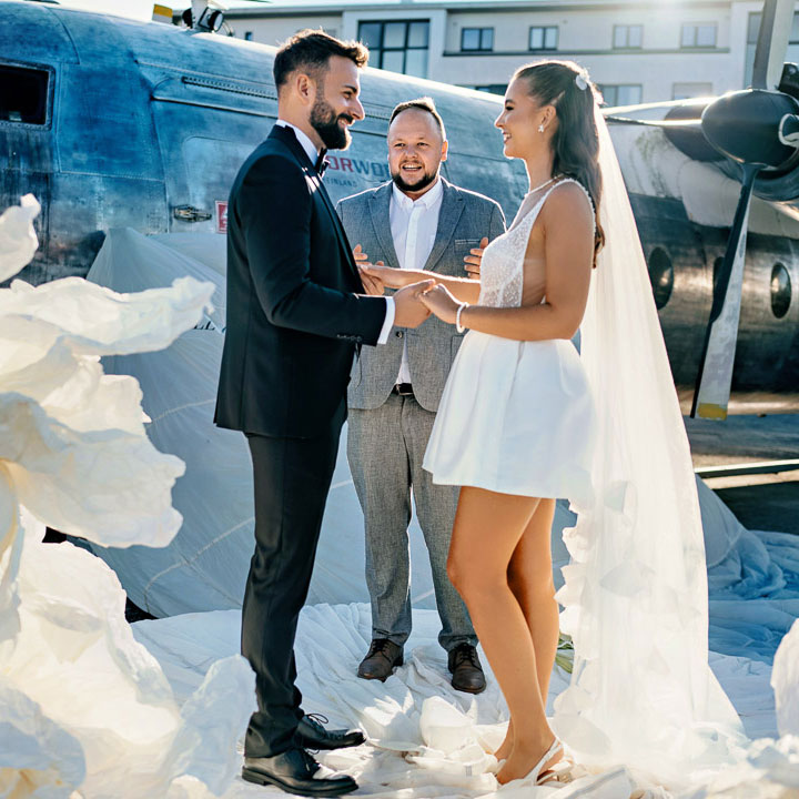 Romantisches Brautpaarfoto mit professionellem Tamada vor Vintage-Flugzeug, kunstvoll arrangierter wehender Schleier sorgt für spektakuläre Aufnahme